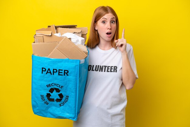 Young redhead woman holding a recycling bag full of paper to recycle isolated on yellow background intending to realizes the solution while lifting a finger up