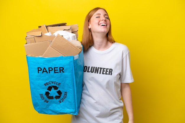 笑いながら黄色の背景に分離されたリサイクルする紙でいっぱいのリサイクルバッグを保持している若い赤毛の女性