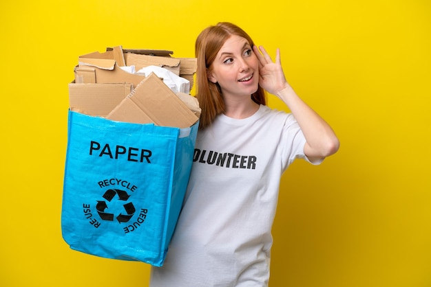 Young redhead woman holding a recycling bag full of paper to recycle isolated on yellow background listening to something by putting hand on the ear