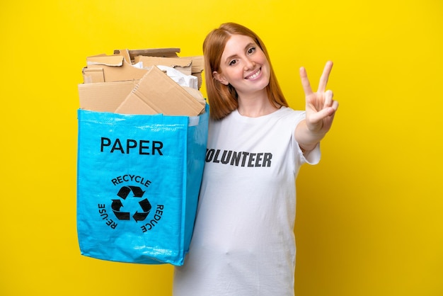 笑顔と勝利のサインを示す黄色の背景に分離された紙でいっぱいのリサイクル バッグを保持している若い赤毛の女性