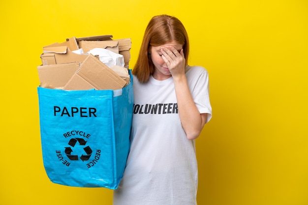 Молодая рыжеволосая женщина, держащая мешок для переработки, полный бумаги для переработки, изолирована на желтом фоне с усталым и больным выражением лица
