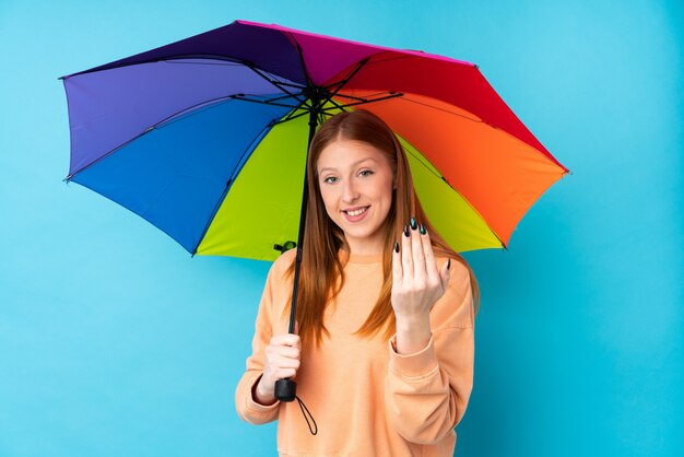 Young redhead woman holding an umbrella over isolated wall inviting to come with hand. Happy that you came