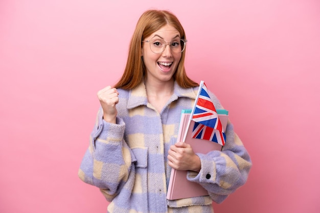 Young redhead woman holding an United Kingdom flag isolated on pink background celebrating a victory in winner position