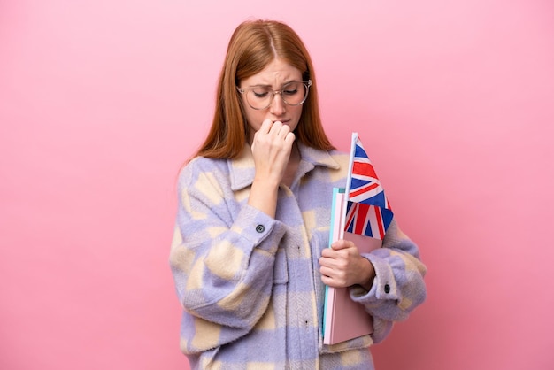 Young redhead woman holding an United Kingdom flag isolated on pink background having doubts