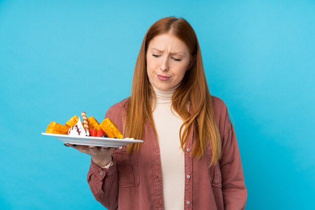 Young redhead woman holding waffles over isolated wall with sad expression