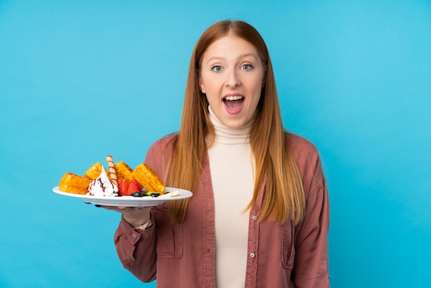 Young redhead woman holding waffles over isolated wall with surprise and shocked facial expression