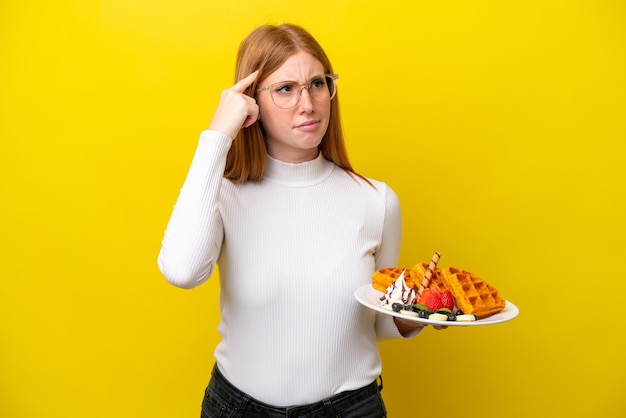 Young redhead woman holding waffles isolated on yellow background having doubts and thinking