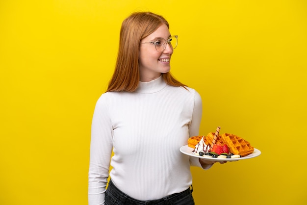 Young redhead woman holding waffles isolated on yellow background looking to the side and smiling