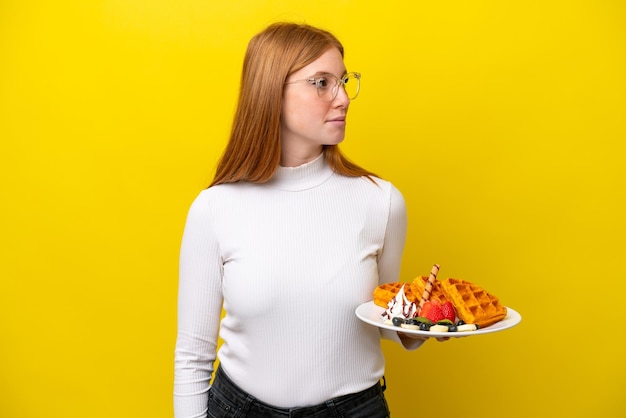 Young redhead woman holding waffles isolated on yellow background looking to the side