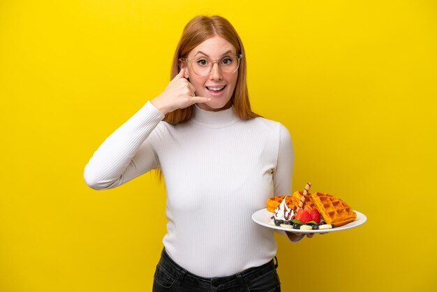 Young redhead woman holding waffles isolated on yellow background making phone gesture call me back sign