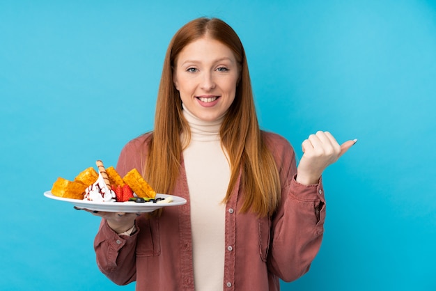 Young redhead woman holding waffles pointing to the side to present a product