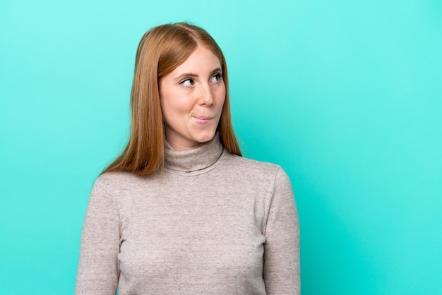 Young redhead woman isolated on blue background having doubts while looking up