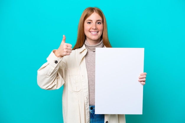 Young redhead woman isolated on blue background holding an empty placard with thumb up