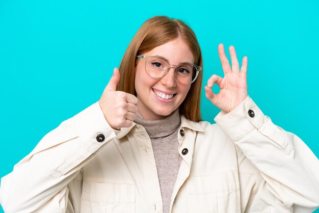 Young redhead woman isolated on blue background with glasses and doing ok sign