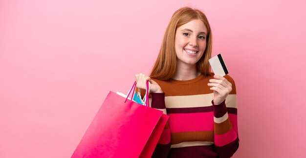 Young redhead woman isolated on pink background holding shopping bags and a credit card