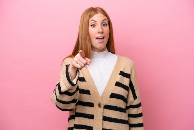 Young redhead woman isolated on pink background surprised and pointing front
