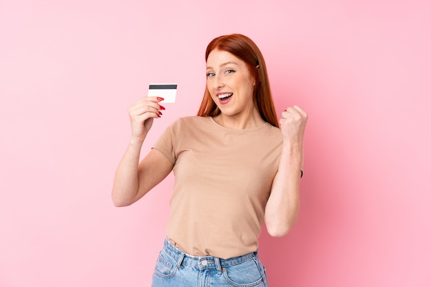 Giovane donna della testarossa sopra il rosa isolato che tiene una carta di credito