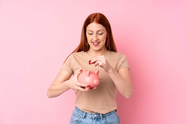 Young redhead woman over isolated pink wall holding a big piggybank