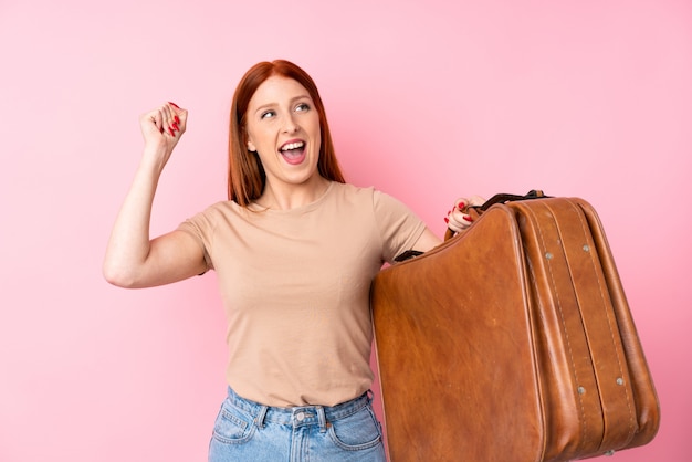 Young redhead woman over isolated pink wall holding a vintage briefcase