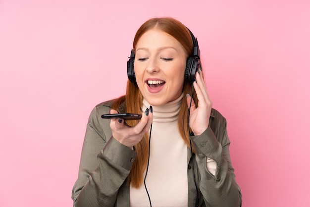 Молодая рыжая женщина над изолированной розовой стеной слушает музыку с мобильного и поет