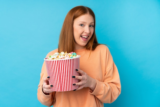 Giovane donna di redhead sopra la parete isolata che tiene un grande secchio di popcorn