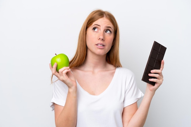 Young redhead woman isolated on white background having doubts while taking a chocolate tablet in one hand and an apple in the other