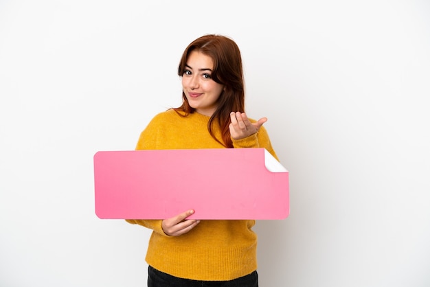 Young redhead woman isolated on white background holding an empty placard and doing coming gesture