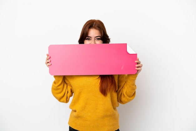 Young redhead woman isolated on white background holding an empty placard and hiding behind it