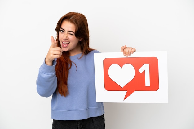 Photo young redhead woman isolated on white background holding a placard with like icon and pointing to the front