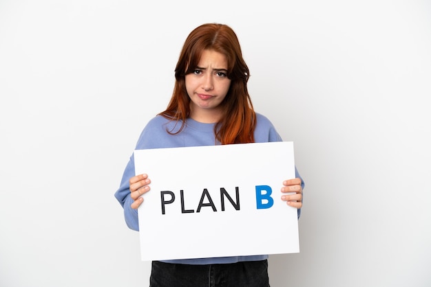 Young redhead woman isolated on white background holding a placard with the message PLAN B with sad expression