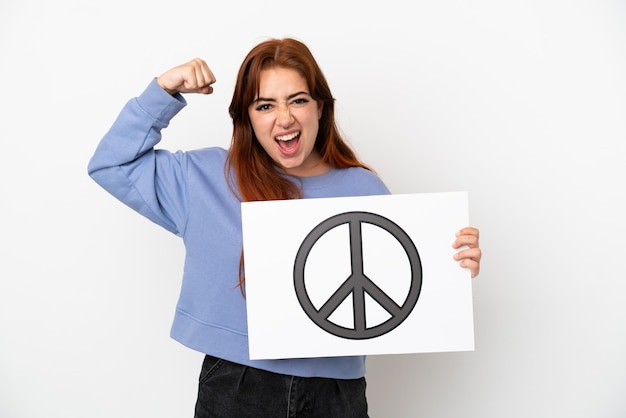 Young redhead woman isolated on white background holding a placard with peace symbol and doing strong gesture