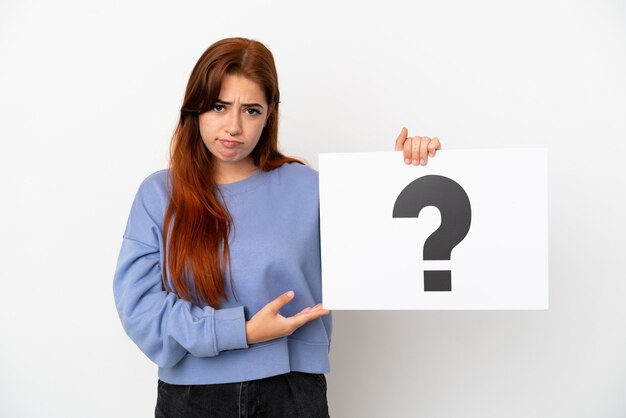 Young redhead woman isolated on white background holding a placard with question mark symbol with sad expression