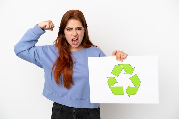 Young redhead woman isolated on white background holding a placard with recycle icon and doing strong gesture