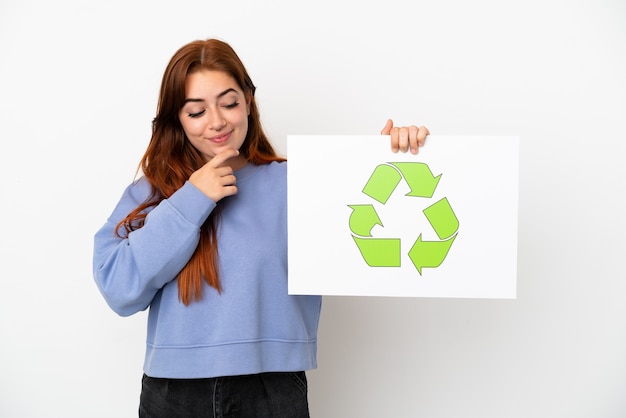 Young redhead woman isolated on white background holding a placard with recycle icon