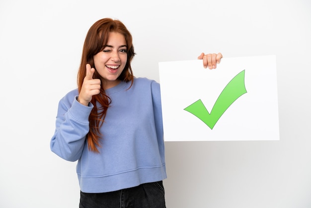 Young redhead woman isolated on white background holding a placard with text Green check mark icon and pointing to the front