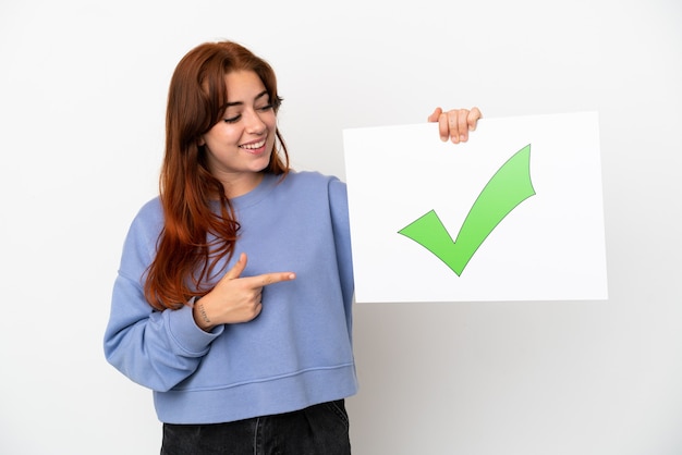 Photo young redhead woman isolated on white background holding a placard with text green check mark icon and  pointing it