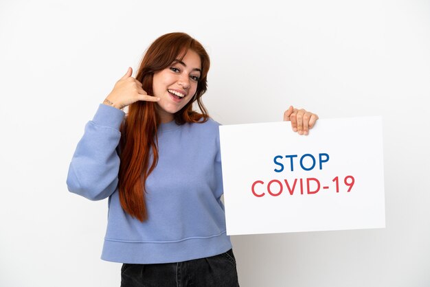 흰색 배경에 격리된 젊은 빨간 머리 여성은 Stop Covid 19라는 문구가 적힌 플래카드를 들고 전화 제스처를 하고 있습니다.