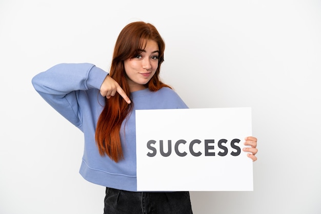 Young redhead woman isolated on white background holding a placard with text SUCCESS and  pointing it