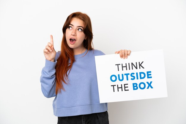 Young redhead woman isolated on white background holding a placard with text Think Outside The Box and thinking