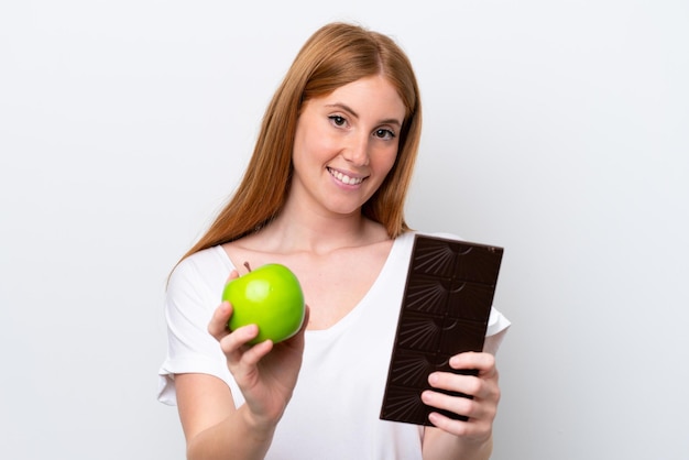 Young redhead woman isolated on white background taking a chocolate tablet in one hand and an apple in the other