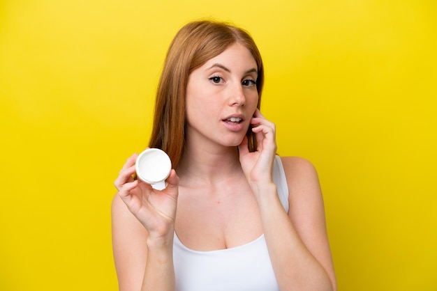 Young redhead woman isolated on yellow background with moisturizer