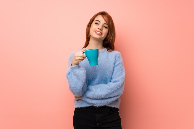 Young redhead woman over pink holding a hot cup of coffee