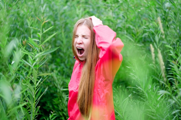 Young redhead woman in pink raincoat yawning due to boring cloudy weather