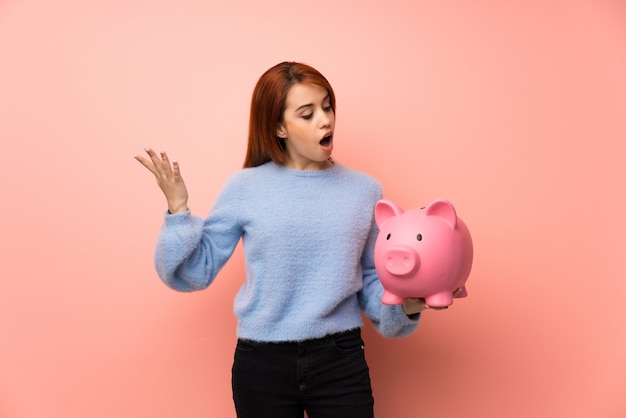 ピンク上の若い赤毛の女性はpiggybankを押しながら驚いた