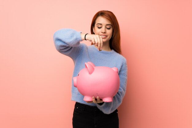 Young redhead woman over pink  taking a piggy bank and happy because it is full