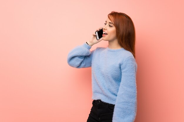 Young redhead woman over pink wall keeping a conversation with the mobile phone