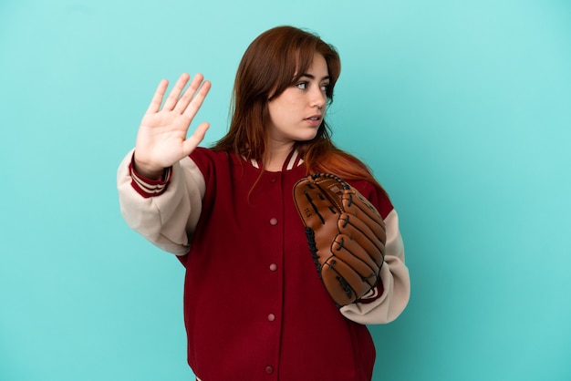 Young redhead woman playing baseball isolated on blue background making stop gesture and disappointed