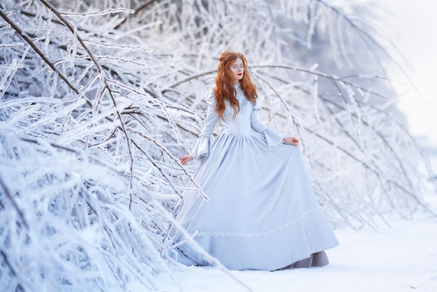 Young redhead woman, a princess, walks in a winter forest in a blue dress. Frost and snow on the trees.