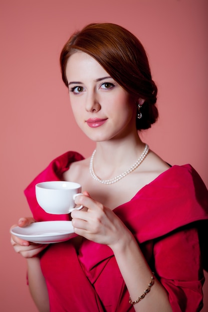 Young redhead woman in red Victorian epoch clothes with cup of tea on pink