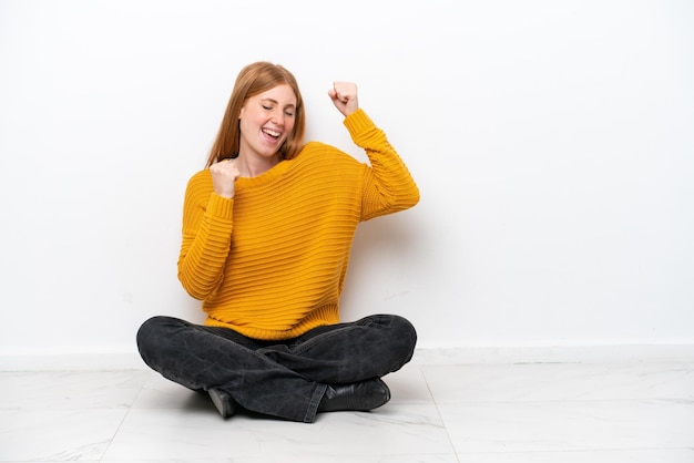 Young redhead woman sitting on the floor isolated on white background celebrating a victory